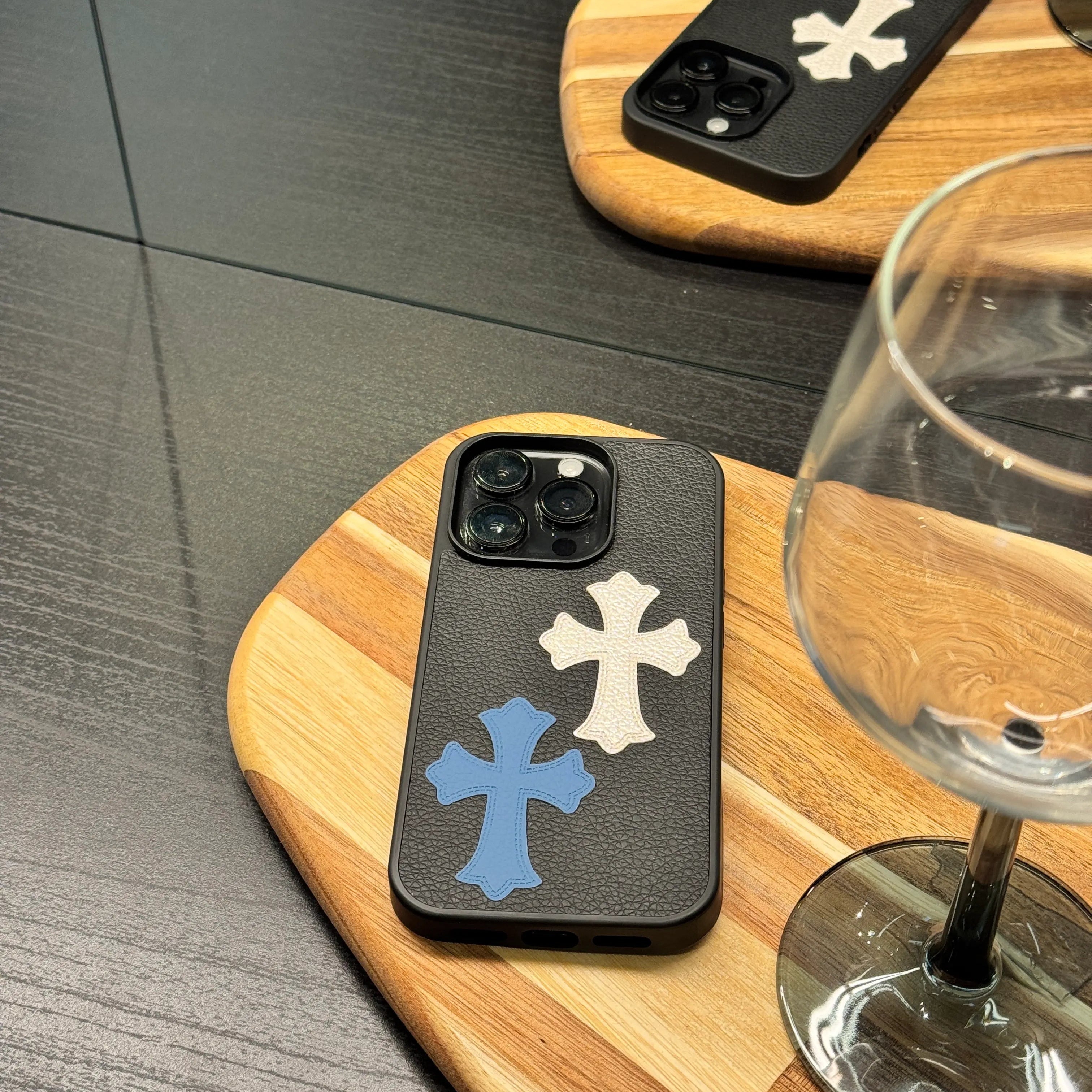 Phone cases with cross designs on a wooden coaster next to a glass of wine.